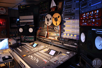 A professional audio recording studio with various mixers, speakers, and reel-to-reel tape machines, surrounded by numerous control panels.